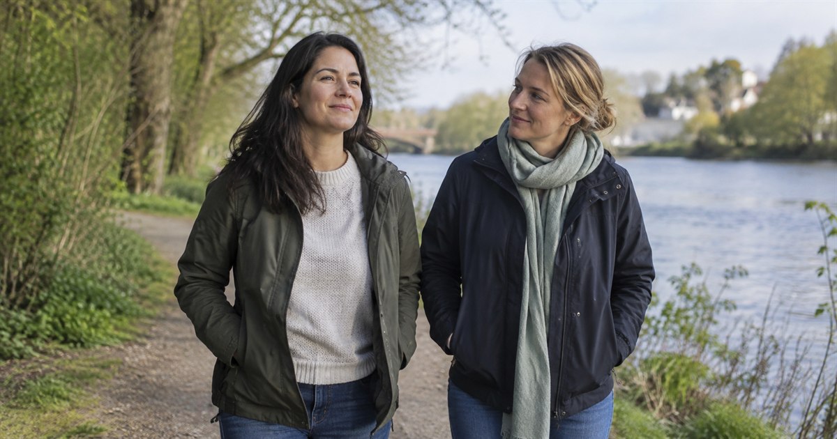 A dignified adult woman taking a steady walk beside a supportive friend on a green riverside path